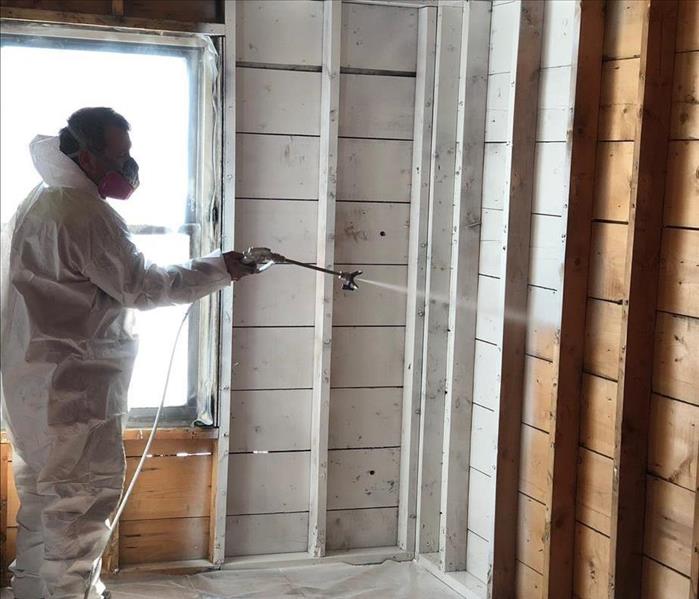 SERVPRO technician in full protective equipment applying paint to corner of deconstructed room with airless sprayer.
