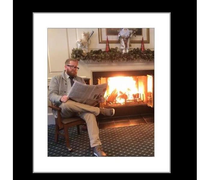 a bearded man wearing glasses sits in a chair by a fireplace while reading a newspaper.