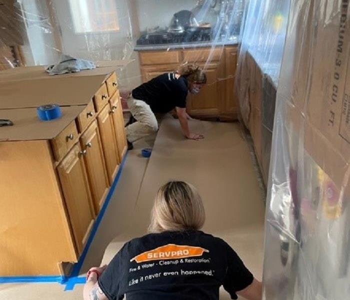 Two SERVPRO technicians carefully placing cardboard on top of kitchen floor. Countertops and cabinets have plastic over them.