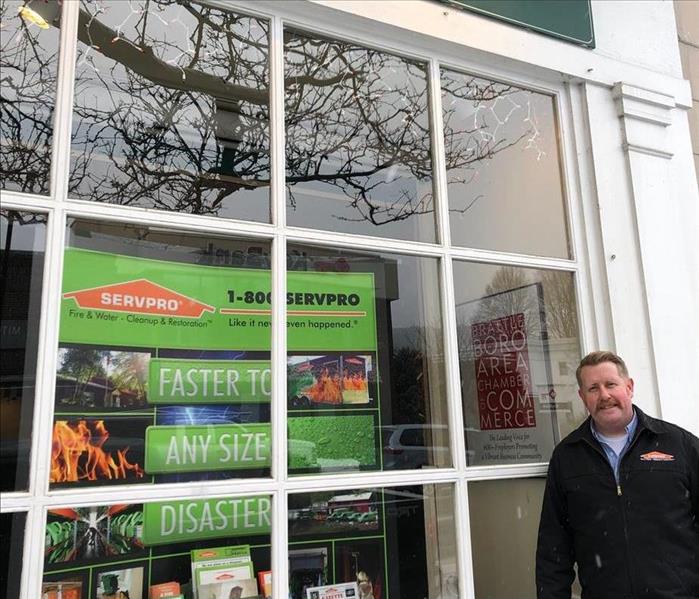 Mustached man wearing a black jacket with SERVPRO logo stands outside a chamber of commerce display window featuring SERVPRO