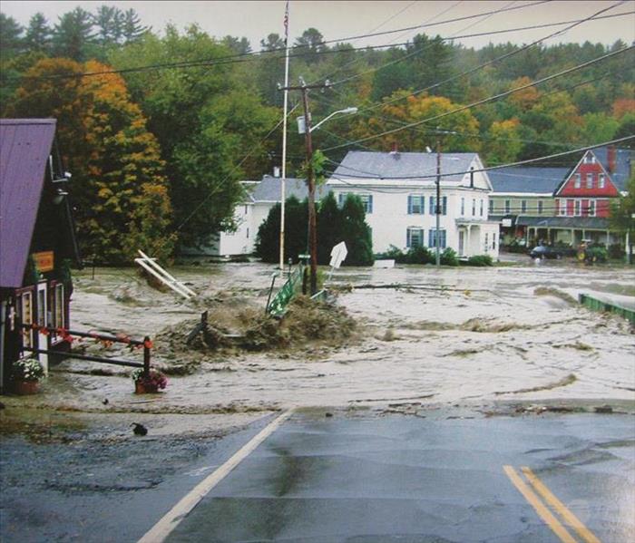 River water has flooded over a main road in a small town causing damage to trees, signs, and property surrounding the road.